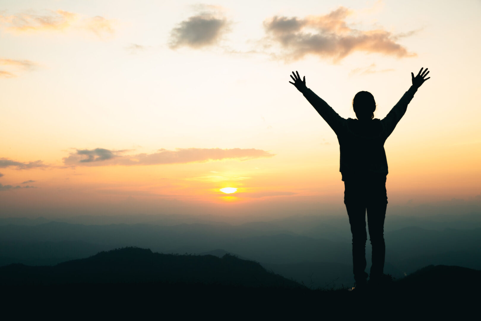 Silhouette of a person with raised arms standing on a hilltop during sunset, with mountains and clouds in the background.