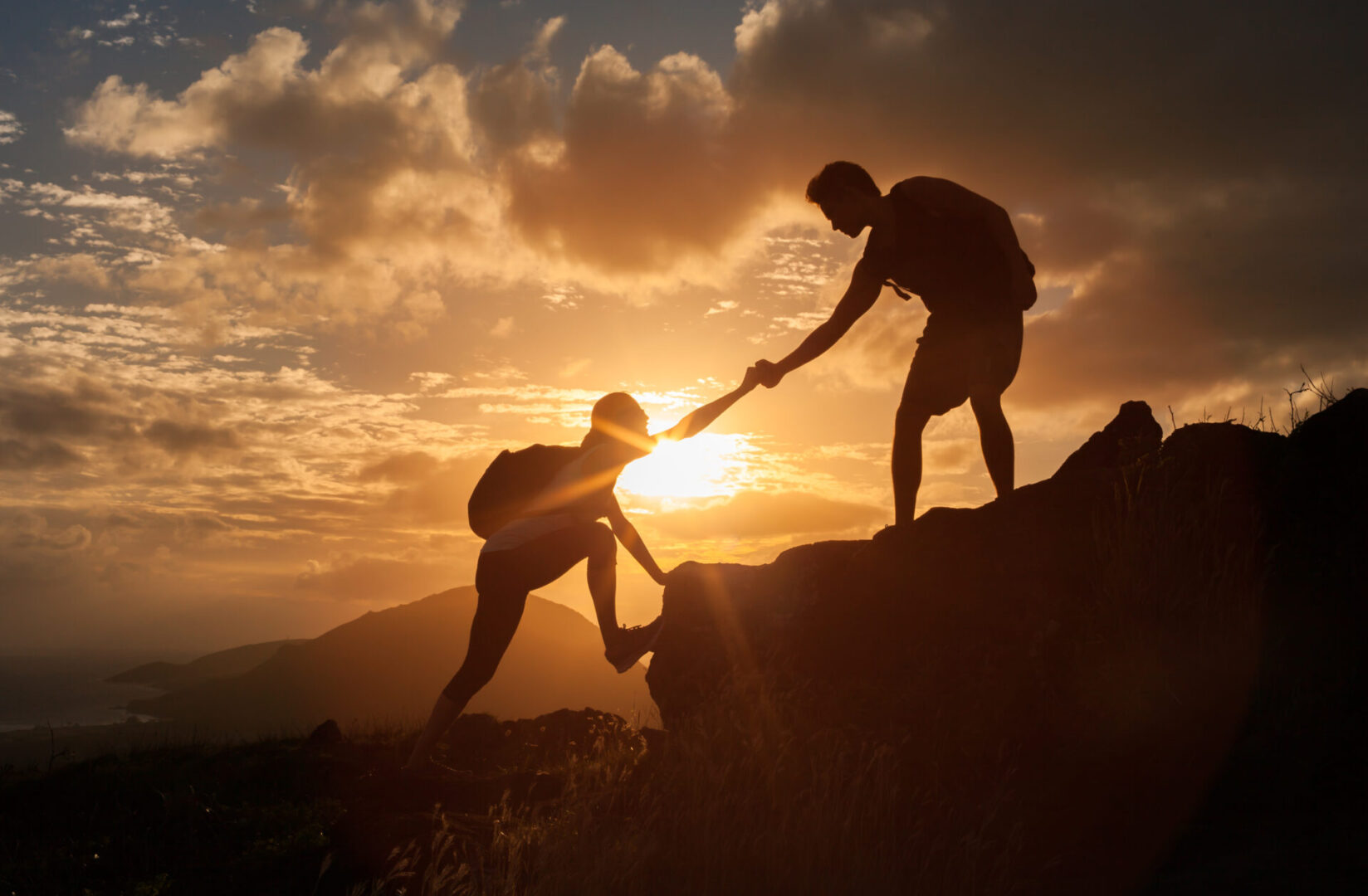 Two silhouetted hikers on a hill at sunset, with one reaching to help the other up.